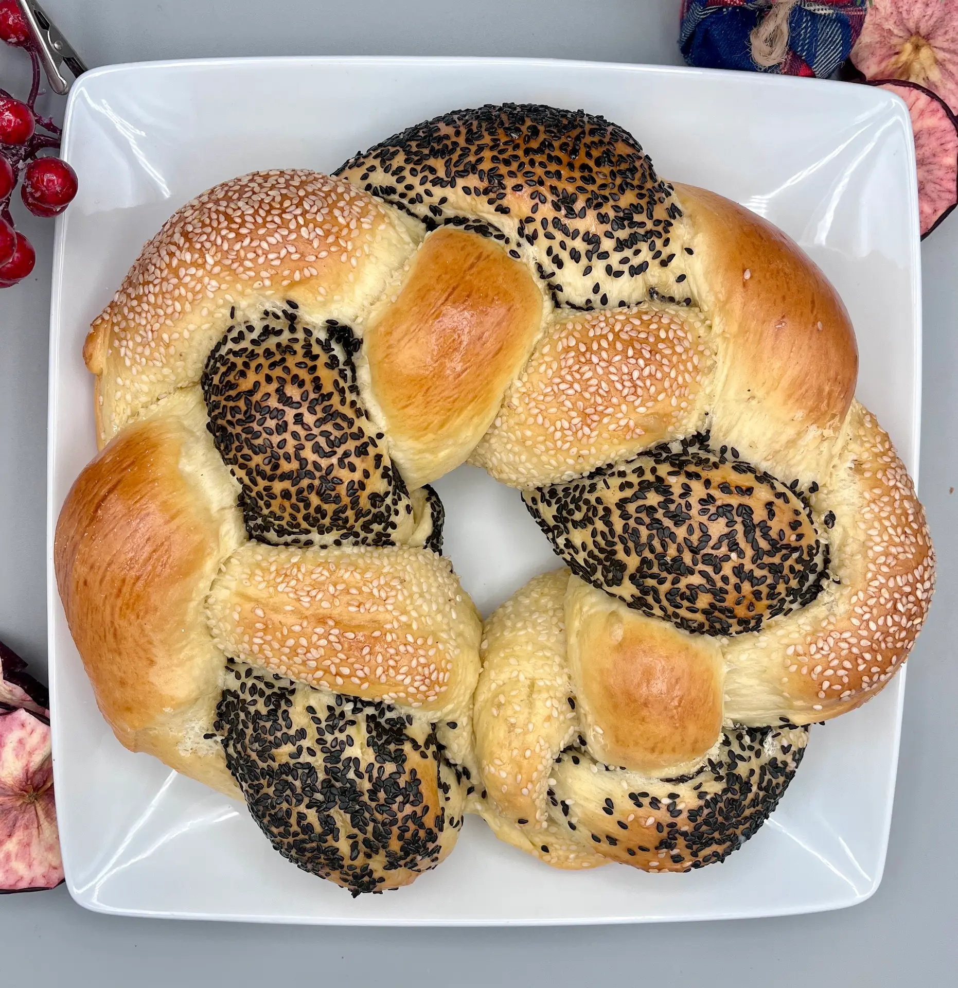 A beautifully braided round challah with black and white sesame seed toppings, displayed on a white platter, symbolizing unity and tradition.